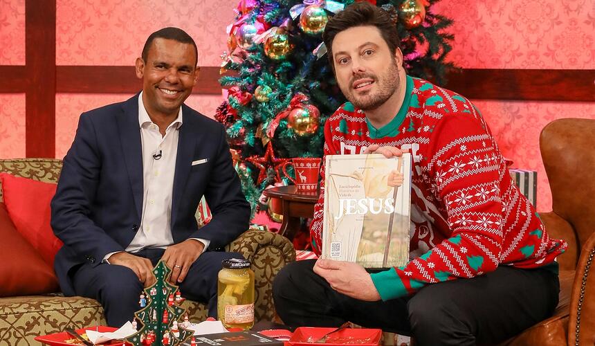 Rodrigo Silva de blazer azul, camisa social branca, calça jeans escura, sentado num sofá ao lado de uma árvore de Natal ao lado de Danilo Gentili, que está sorrindo, sentado, segurando um livro durante gravação do The Noite, do SBT