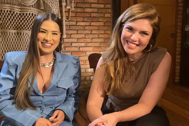 Simone Medes de blazer azul claro, sorrindo, com colar de brilhantes, cabelo liso ao lado de repórter durante gravação de entrevista para o Fantástico, da TV Globo