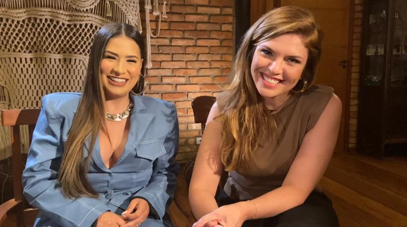 Simone Medes de blazer azul claro, sorrindo, com colar de brilhantes, cabelo liso ao lado de repórter durante gravação de entrevista para o Fantástico, da TV Globo