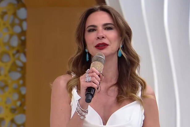 Luciana Gimenez, com uma roupa branca, microfone em mãos, em trecho do SuperPop