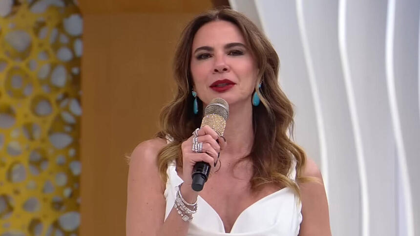 Luciana Gimenez, com uma roupa branca, microfone em mãos, em trecho do SuperPop