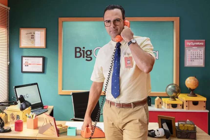 Tadeu Schmidt de calça bege, camisa social branca, gravata azul, segurando um telefone laranja num escritorio durante gravação de chamada para o BBB, da TV Globo