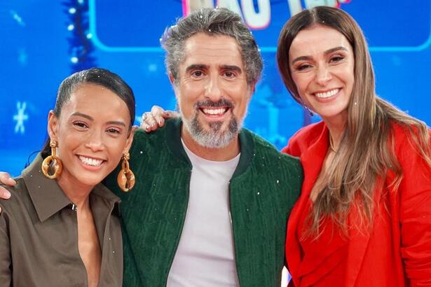 Taís Araujo de jaqueta verde musco, cabelo preso, liso, com brincos dourados sorrindo; Marcos Mion de jaqueta jeans verde, camisa branca, sorrindo; Mônica Martelli de camisa vermelha, cabelo liso pro lado esquerdo, sorrindo durante gravação do Caldeirão, da TV Globo