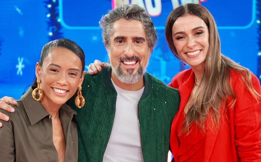 Taís Araujo de jaqueta verde musco, cabelo preso, liso, com brincos dourados sorrindo; Marcos Mion de jaqueta jeans verde, camisa branca, sorrindo; Mônica Martelli de camisa vermelha, cabelo liso pro lado esquerdo, sorrindo durante gravação do Caldeirão, da TV Globo