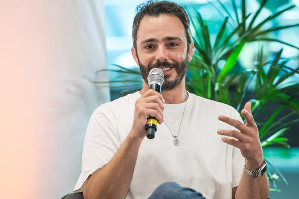 Thiago Rodrigues, com uma blusa branca e microfone na mão, sentado em trecho da coletiva de Valor da Vida, na Band