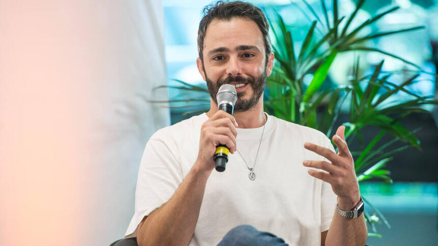 Thiago Rodrigues, com uma blusa branca e microfone na mão, sentado em trecho da coletiva de Valor da Vida, na Band