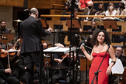 Dora Morelenbaum de macaquito vermelho, segurando o microfone no teatro Sala São Paulo em frente a orquestra durante gravação do tributo a Gal Costa