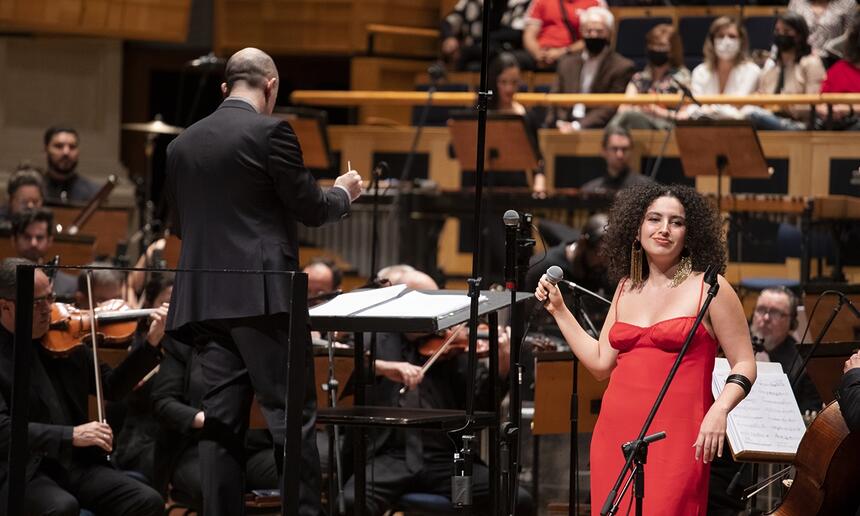 Dora Morelenbaum de macaquito vermelho, segurando o microfone no teatro Sala São Paulo em frente a orquestra durante gravação do tributo a Gal Costa