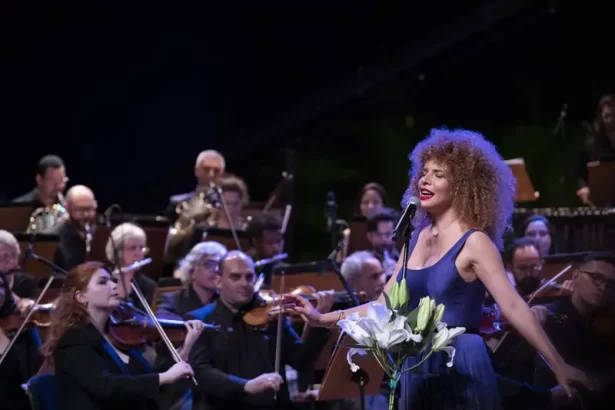 Vanessa da Mata com vetido azul, braços abertos, batom vermelho, cabelo volumoso, solto, durante apresentação