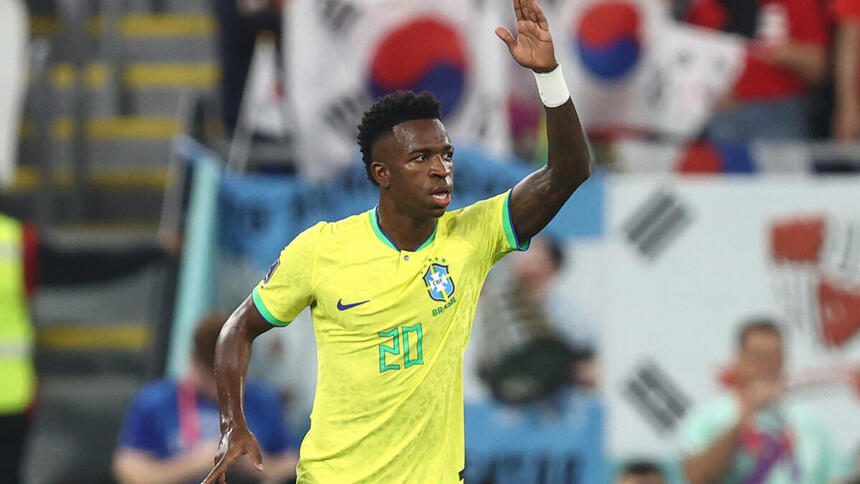 Vinícius Jr, em campo com a camisa do Brasil, com a mão esquerda levantada