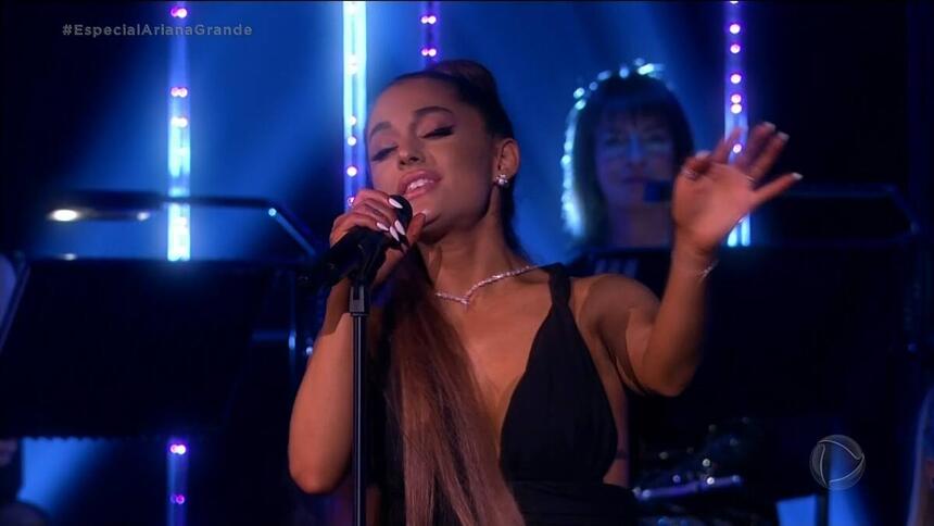 Ariana Grande levanta o braço esquerdo para canta no especial Live in London, exibido na Record TV