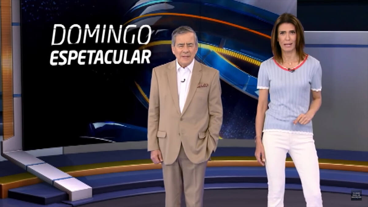 Paulo Henrique Amorim e Janine Borba no cenário virtual do Domingo Espetacular. Os apresentadores estão em pé na frente do telão que exibe o novo logo em flat do Domingo Espetacular