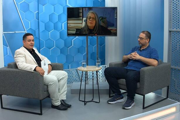 Apresentador do Aparecida Debate vestido de roupa branca e camisa preta junto com médico convidado vestido de camisa azul e calça preta. Na tela do estúdio do programa tem também outra convidada do programa.