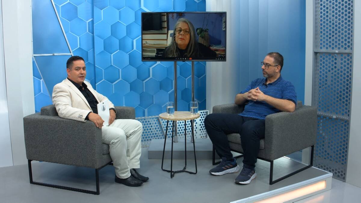 Apresentador do Aparecida Debate vestido de roupa branca e camisa preta junto com médico convidado vestido de camisa azul e calça preta. Na tela do estúdio do programa tem também outra convidada do programa.