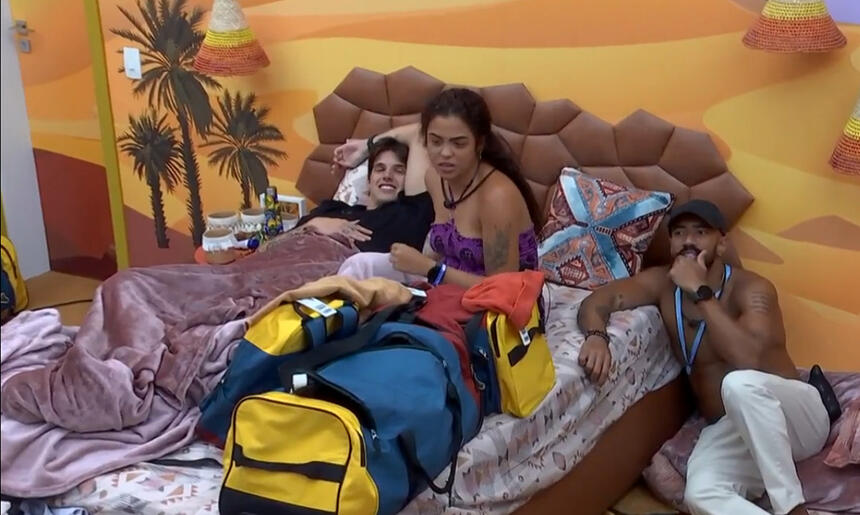 Gabriel Fop de camisa preta, deitado ao lado de Paula Freitas, de top roxo, ao lado de Ricardo de camisa preta e calça branca sentado no chão do Quarto Deserto do BBB 23