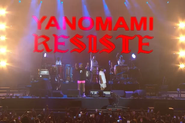 Criolo com short rosa, camisa preta, punho cerrado ao lado da banda e um telão escrito Yanomamis resiste