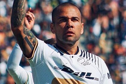 Daniel Alves no campo de futebol vestindo uma camisa branca com detalhes e com semblante sério.