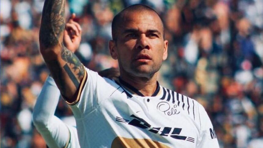 Daniel Alves no campo de futebol vestindo uma camisa branca com detalhes e com semblante sério.