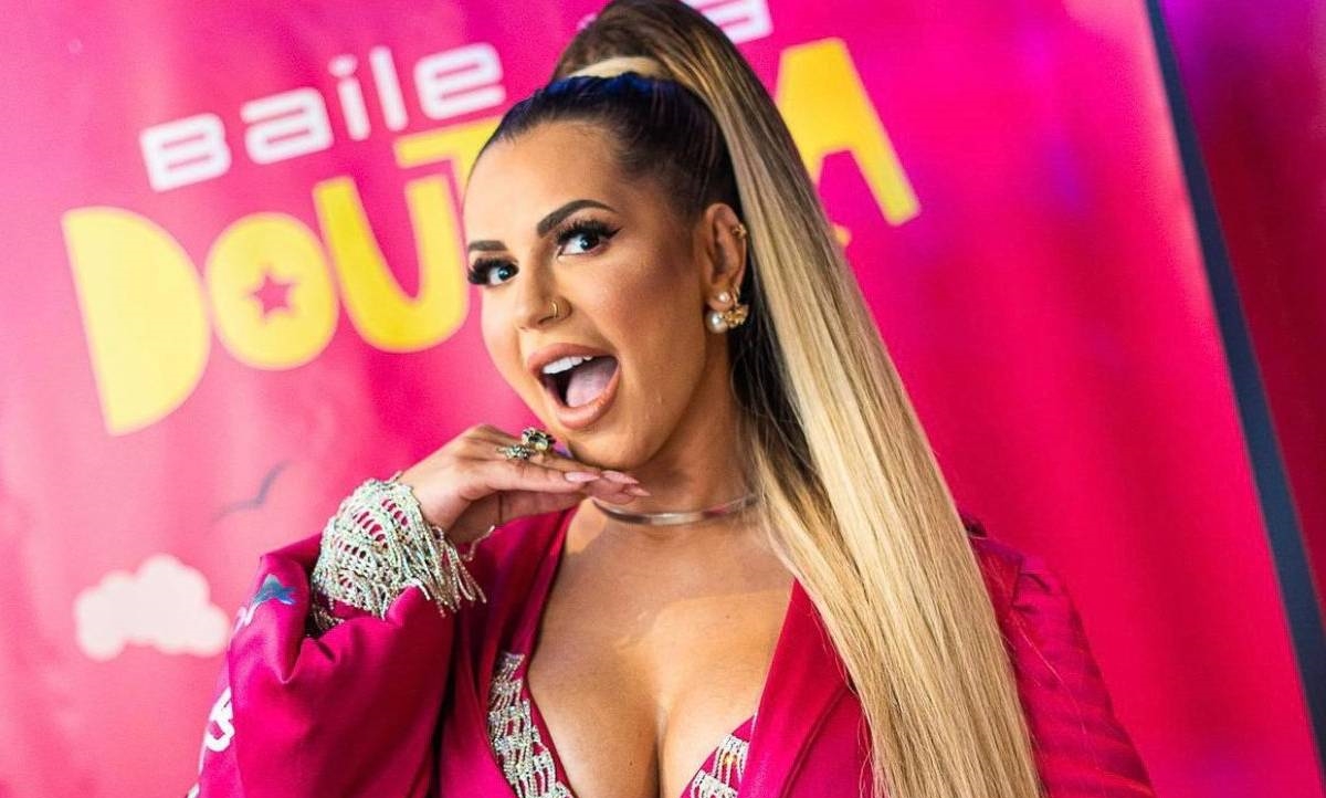 Deolane Bezerra com a mão no queixo, boca aberta, usando blazer decotado na cor rosa durante presença vip em festa