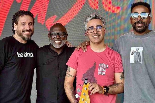 Duca Tambasco de camisa em tom de magenta, segurando uma guitarra, de óculos de grau, sorrindo, ao lado de Kleber Lucas, de camisa preta, sorrindo, de óculos de grau, ao lado de Leonardo Gonçalves de camisa preta, sorrindo, usando barba e Clovis Damásio de camisa cinza, com a mão no ombro de Duca