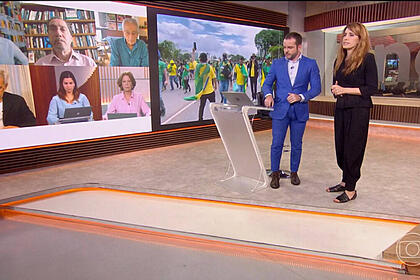Erick Bang e Poliana Abritta, em pé nos estúdios da GloboNews, durante a cobertura dos atos terroristas. Comentaristas aparecerem no telão