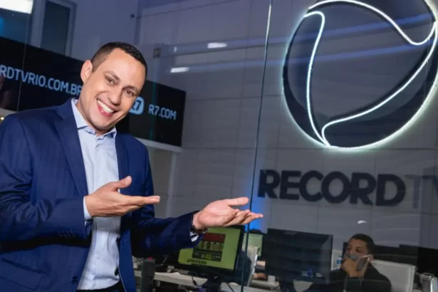 Ernani Alves na redação de jornalismo da Record TV Rio, apontando para o logo