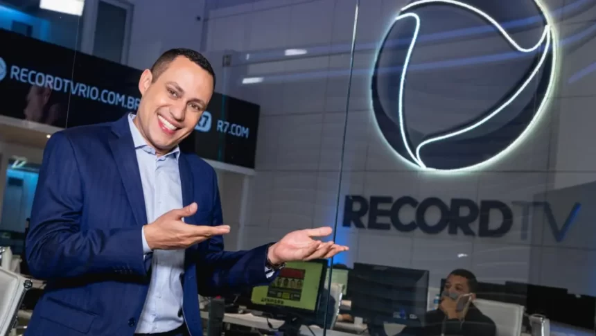 Ernani Alves na redação de jornalismo da Record TV Rio, apontando para o logo