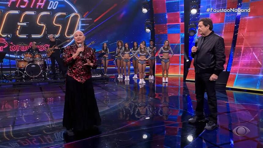 Fafá de Belém vestida de vermelho e preto segurando o microfone ao lado de Fausto Silva vestido de roupa preta no palco do Faustão Na Band.