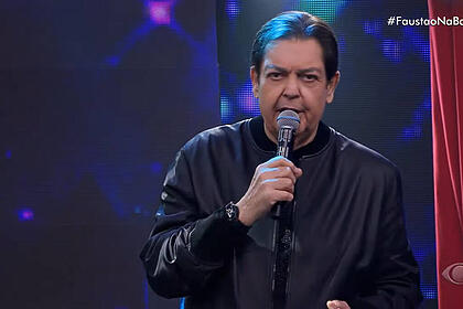 Fausto Silva vestido com jaqueta preta no palco do Faustão Na Band segundo o microfone no palco do seu programa.