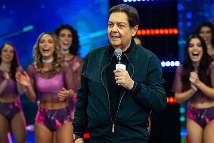 Fausto Silva no palco do seu programa na Band, vestido uma camisa preta e jaqueta verde, sorrindo ele segura seu microfone. Ao fundo também sorrindo e vestindo roupas roxas, está suas bailarinas.