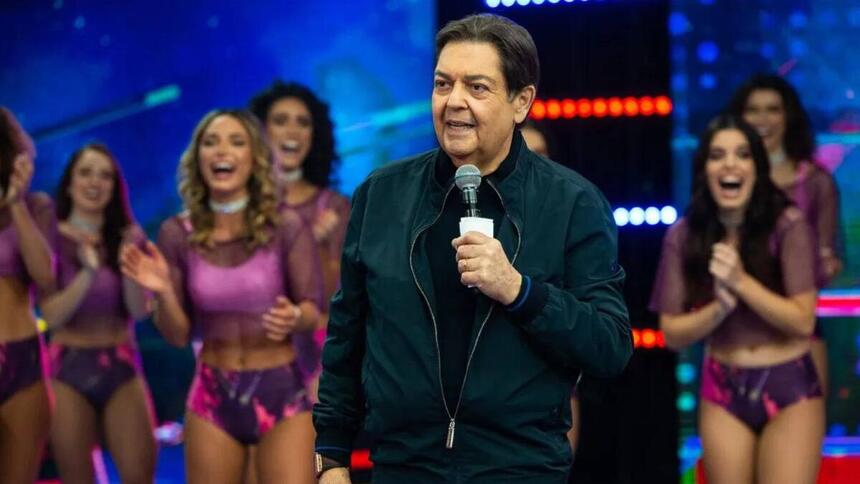 Fausto Silva no palco do seu programa na Band, vestido uma camisa preta e jaqueta verde, sorrindo ele segura seu microfone. Ao fundo também sorrindo e vestindo roupas roxas, está suas bailarinas.