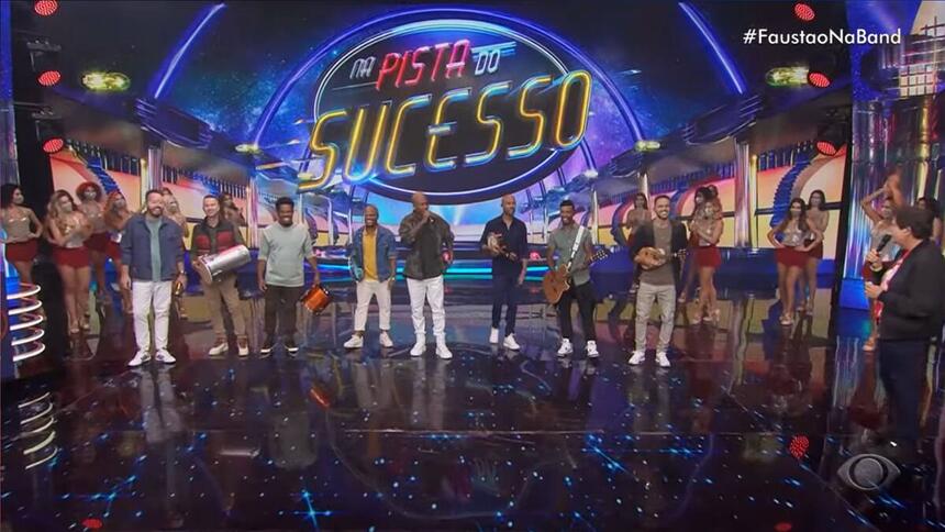 Palco do Faustão Na Band com a Turma do Pagode no início de sua apresentação musical. No canto do palco está Fausto Silva com roupa escura.