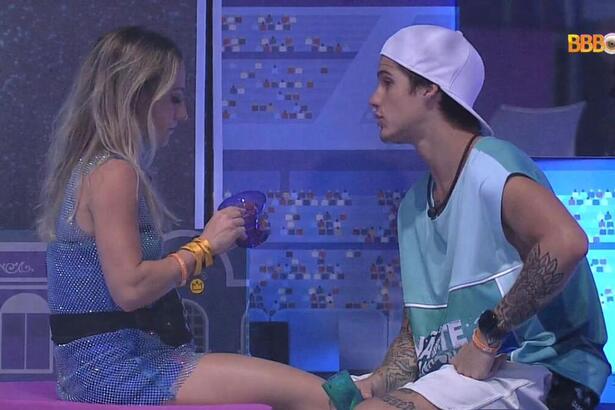 Bruno sentada segurando um copo de roupa brilhosa de frente para Gabriel que está sentado vestido com uma camisa azul e bermuda branca.