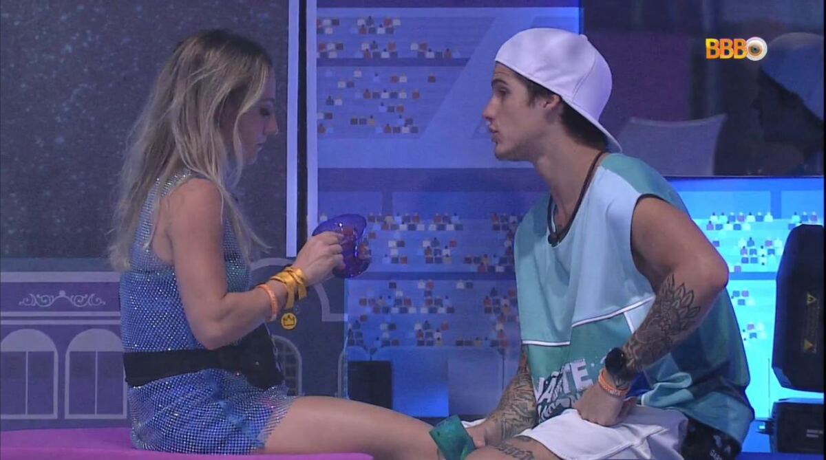 Bruno sentada segurando um copo de roupa brilhosa de frente para Gabriel que está sentado vestido com uma camisa azul e bermuda branca.