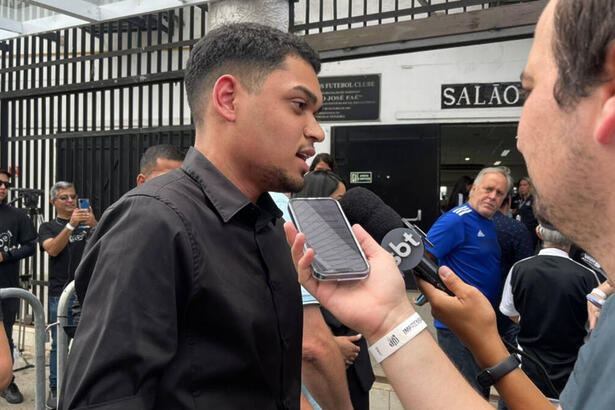 Gabriel, neto do Pelé, vestido de preto, sendo entrevistado por jornalistas na porta da Vila Belmiro