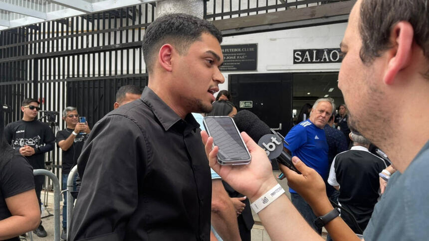 Gabriel, neto do Pelé, vestido de preto, sendo entrevistado por jornalistas na porta da Vila Belmiro