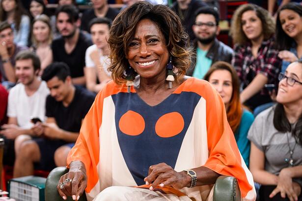 Gloria Maria sorrindo, com camisa laranja com estampa azul e bege, sentada durante participação no Altas Horas