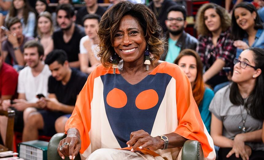 Gloria Maria sorrindo, com camisa laranja com estampa azul e bege, sentada durante participação no Altas Horas