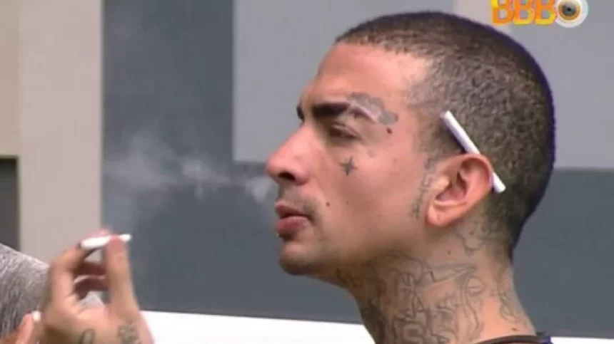 MC Guimê fumando, sentado, dentro da casa do BBB 23, com cigarro na orelha, soltando fumaça pelo nariz