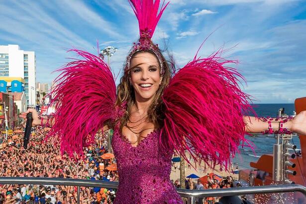 Ivete Sangalo sorrindo, com os braços abertos, com look rosa e plumas durante performance em trio no Carnaval de Salvador