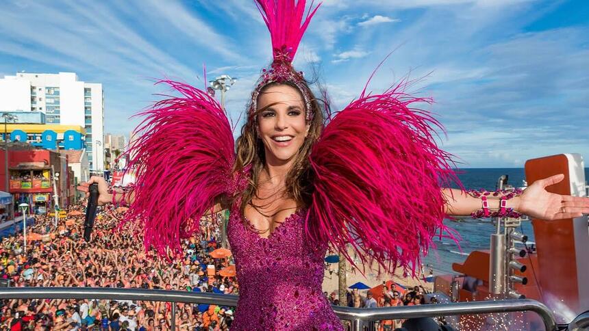 Ivete Sangalo sorrindo, com os braços abertos, com look rosa e plumas durante performance em trio no Carnaval de Salvador