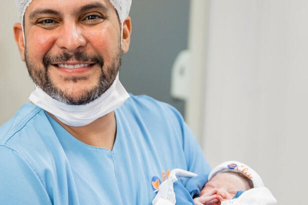 Jorge com a roupa de centro cirúrgico, com a filha recém-nascida no colo