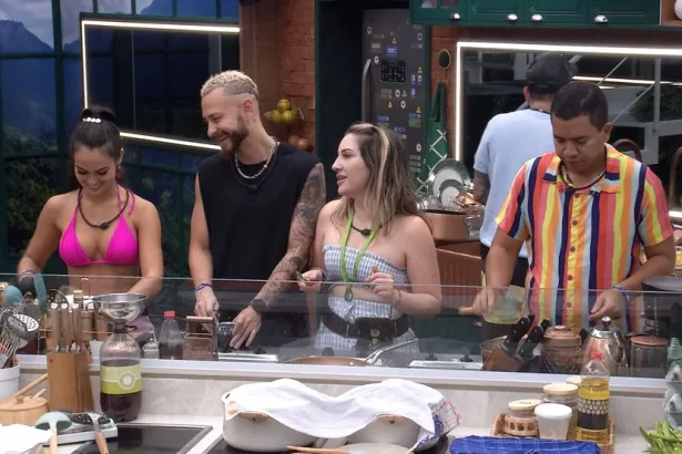 Fred de camisa preta, com a cabeça de lado, sorrindo, cortando algo; Amanda de top e saia brancos com linhas azuis olhando pra Larissa; Larissa de camisa rosa, cabelo preso; Bruno de camisa listrada colorida cozinhando