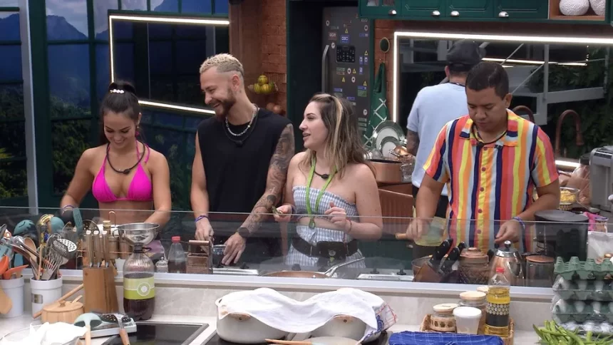 Fred de camisa preta, com a cabeça de lado, sorrindo, cortando algo; Amanda de top e saia brancos com linhas azuis olhando pra Larissa; Larissa de camisa rosa, cabelo preso; Bruno de camisa listrada colorida cozinhando