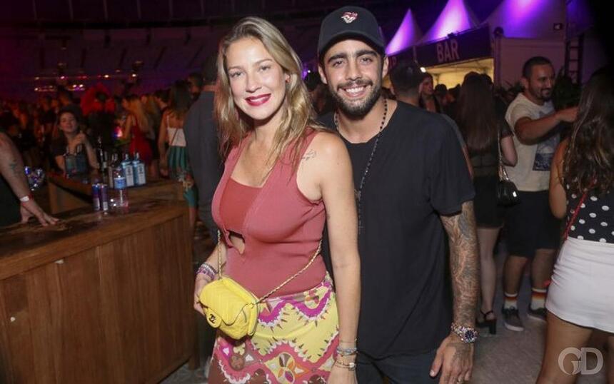 Luana Piovani de camisa em tom de magenta, saia estampada, segurando uma bolsa amarela, sorrindo, abraçada por Pedro Scooby, que está de chapéu e camisa preta