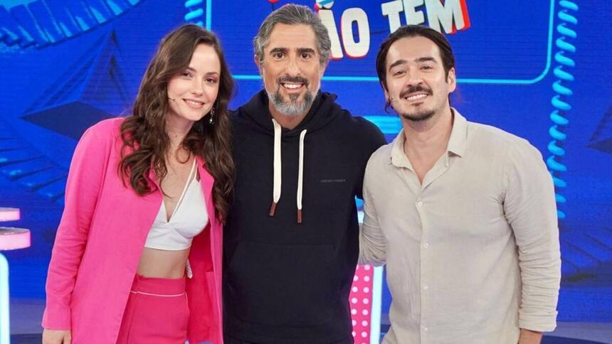 Marcos Mion vestido com roupa preta no meio dos seus convidados. Com ele está Marcos Veras vestido com roupa branca e Rosanne Mulholland com roupa rosa.