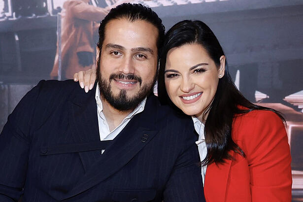 Maite Perroni de camisa vermelha e branca, sorrindo ao lado do marido Andrés Tovar, abraçados durante gravação de filme para Netflix