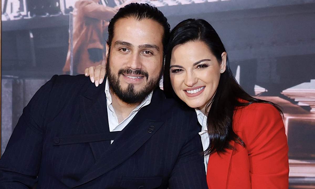 Maite Perroni de camisa vermelha e branca, sorrindo ao lado do marido Andrés Tovar, abraçados durante gravação de filme para Netflix
