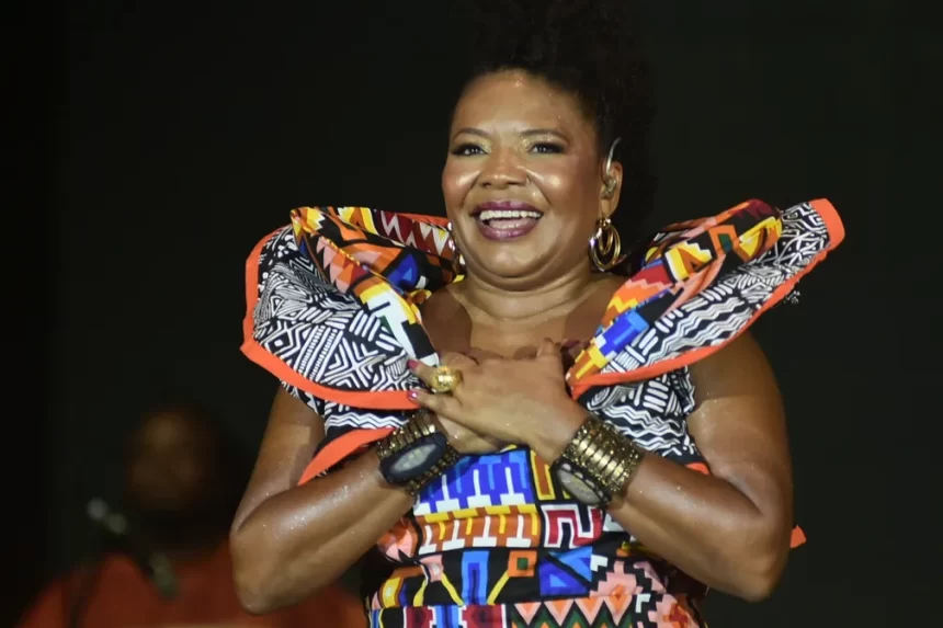 Margareth Menezes com as mãos no peito, sorrindo, usando roupa futuristica em tons de magenta, ciano e branco durante show no Festival de Verão de Salvador