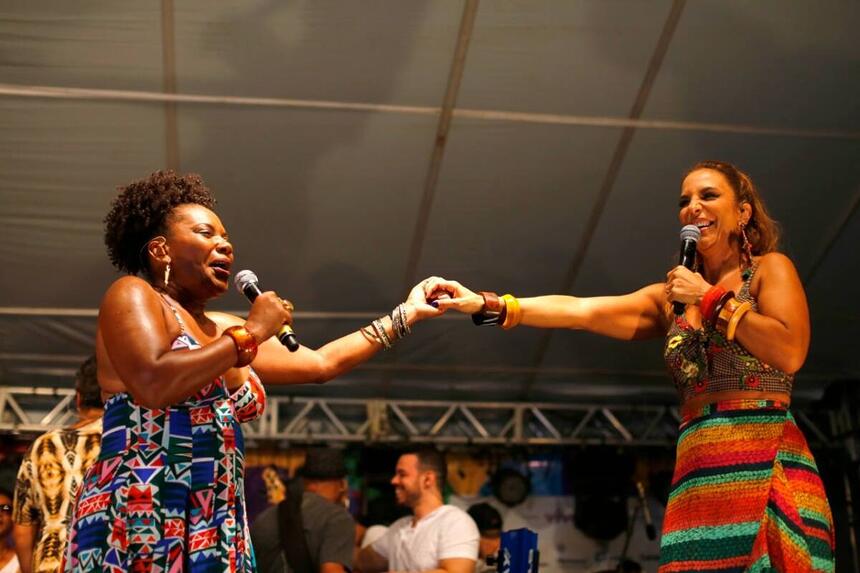 Margareth Menezes de vestido estampado em tons de ciano, segurando a mão de Ivete Sangalo durante show em Salvador